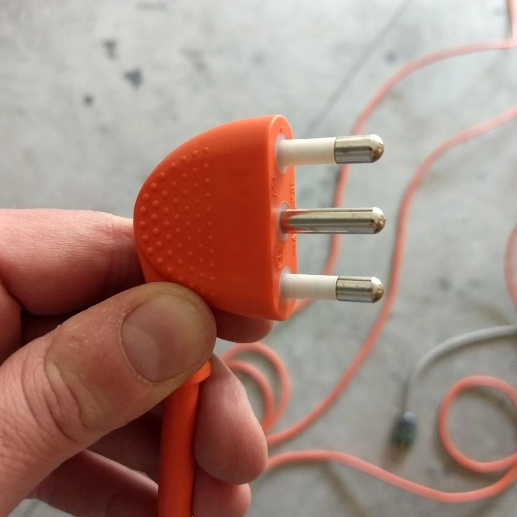 Una mano que sostiene el enchufe de un alargador naranja, con el cable desenrollado por el suelo del patio