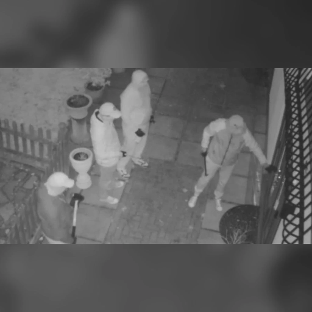 Imagen nocturna por infrarrojos de una cámara: cuatro personas encapuchadas en el patio de una casa, una intenta abrir la puerta